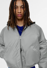 Grijze bomberjack met ritssluiting, doorgestikte details en een zwarte capuchon. Gladde textuur, geribbelde boorden en zijzakken. Blauwe blouse eronder.