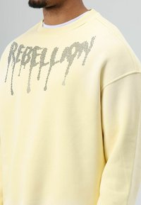Lichtgele sweatshirt met een ontspannen pasvorm, voorzien van een druppelend tekstontwerp in zilveren versiering over de voorkant.