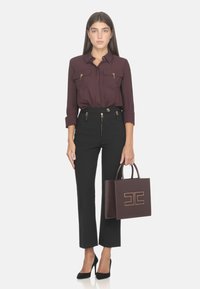 Blusa bordeaux con due tasche sul petto, pantaloni neri sartoriali con zip dorate e una borsa tote strutturata color marrone scuro con logo dorato.
