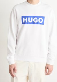 Biała bluza z długim rękawem, z dużą niebieską prostokątną grafiką z napisem "HUGO" w białym kolorze. Rybkowane mankiety i dół.