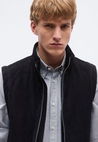 Fekete szarvasbőr mellény magas nyakrésszel, cipzáras elejű, halványkék gombos ingre viselve. Tiszta design, klasszikus szabás.