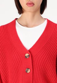 Rode gebreide cardigan met een V-halslijn, voorzien van grote knopen en een getextureerd patroon. Draagt over een wit shirt.
