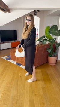 Svart lang kjole med lange ermer, beige slip-on sko, hvit veske med brune detaljer. Rom med tregulv, grønn plante og oransje sofa.