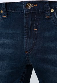 Jeans de mezclilla azul oscuro con cremallera y cierre de botón. Presentan costuras naranjas en contraste y un botón de marca en la cintura.