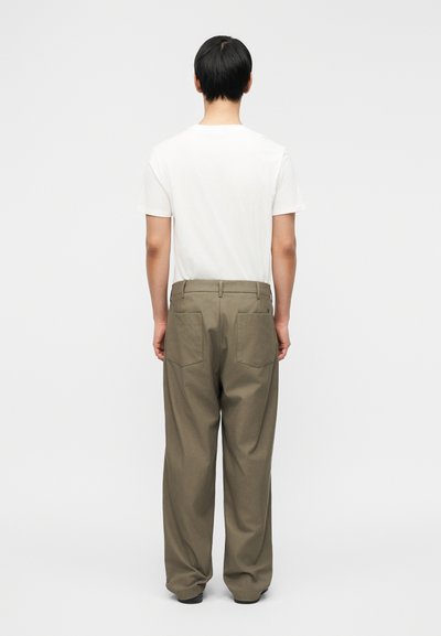 Homme de dos portant une chemise blanche à manches courtes, un pantalon ample vert olive avec des poches arrière, et des chaussures noires, sur un fond uni.