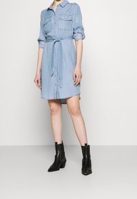 Ljust blå denimskjolklänning med knappar framtill, två bröstfickor och bälte i midjan; matchad med svarta ankelboots.