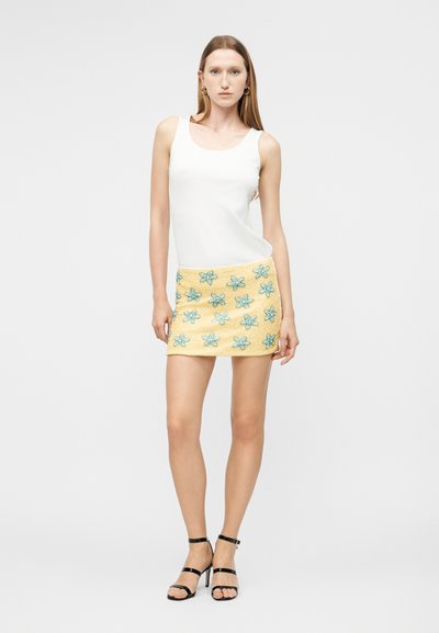 Γυναίκα όρθια με λευκό αμάνικο τοπ, κίτρινη μίνι φούστα με μπλε ανθόμενα σχέδια και μαύρα πέδιλα με ψηλό τακούνι, μπροστά σε απλό φόντο.