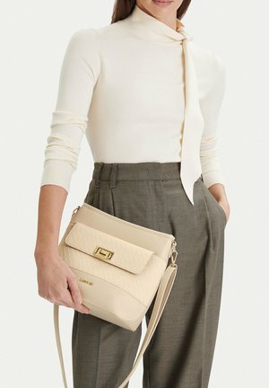 Donna che indossa una maglia crema a maniche lunghe e pantaloni grigi, che tiene una borsa a tracolla beige con patta testurizzata e fermaglio dorato.
