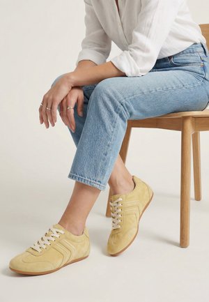 Osoba ubrana w jasnoniebieskie dżinsy i beżowe zamszowe trampki, siedząca na drewnianym krześle, z rękami spoczywającymi na kolanie na jednolitym tle.