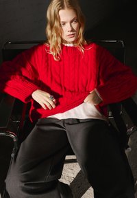 Maglione rosso a maglia intrecciata con vestibilità ampia, polsini a costine e una camicia bianca sovrapposta. Abbinato a pantaloni larghi e scuri.