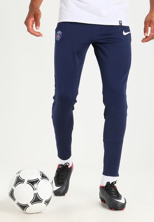 Pantalons de sport navy avec une coupe ajustée, arborant un logo PSG sur la cuisse et le swoosh blanc de Nike. Portés avec des chaussures de foot noires et rouges.