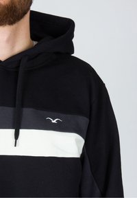 Hombre con hoodie negro con una franja horizontal gris y blanca y un pequeño logo de gaviota blanca en el pecho, contra un fondo claro y liso.