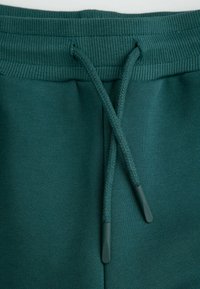 Zielone dresy z prążkowanym pasem, sznurkiem z teksturowanymi końcówkami oraz gładką powierzchnią materiału.