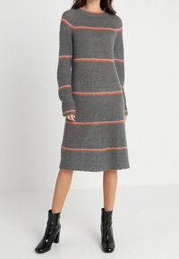 Grå stickad klänning med långa ärmar, med horisontella orangea ränder. Hög rund halsringning och rak passform, bärs med svarta ankelstövlar.