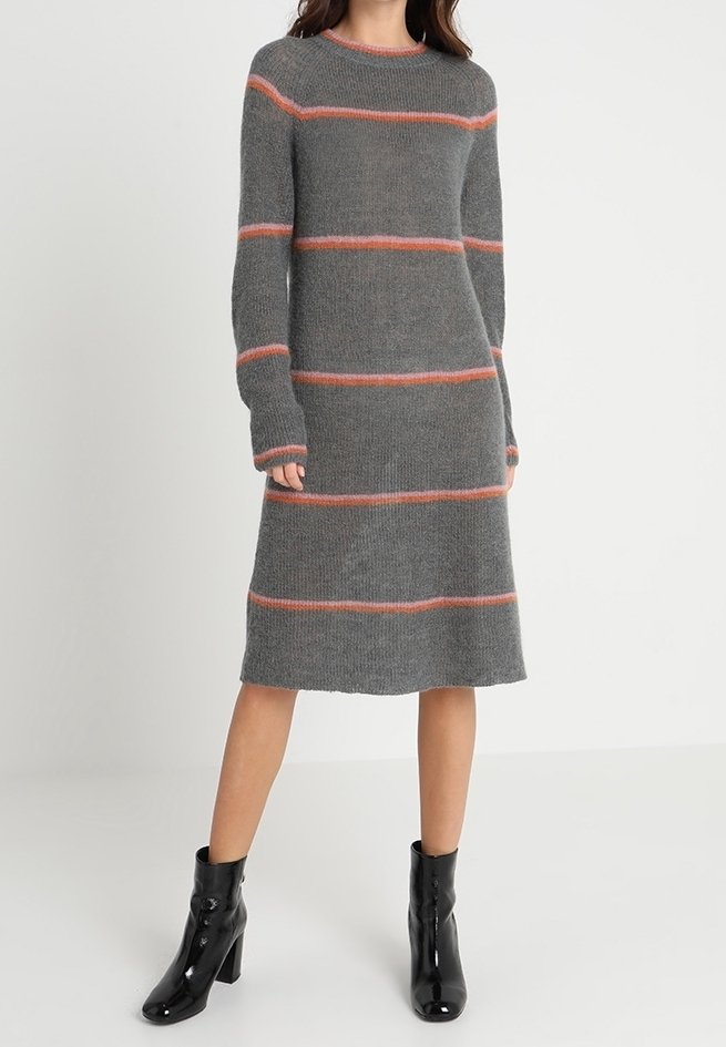 Grå stickad klänning med långa ärmar, med horisontella orangea ränder. Hög rund halsringning och rak passform, bärs med svarta ankelstövlar.