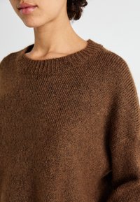 Pull en maille marron avec un col rond, présentant une texture douce et une coupe ample, mettant en valeur le col et les manches côtelés.