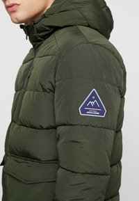 Hombre que lleva una chaqueta acolchada de color verde oscuro con capucha y un parche triangular del logo "Jack & Jones" en la parte superior de la manga.