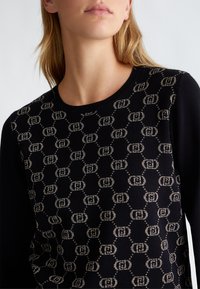 Maglione di maglia nera con un motivo a monogramma dorato, colletto rotondo e maniche a contrasto. Texture morbida con una silhouette aderente.
