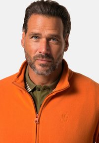 Orange fleece jacket with a zippered front, featuring a stand collar. Underneath is a green collared shirt with light stripes.