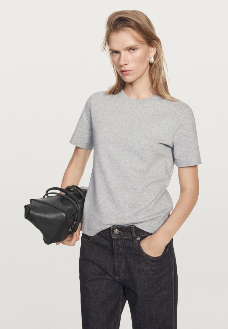 Femme aux cheveux blonds raides portant un t-shirt gris à manches courtes et un jean foncé, tenant un sac à main noir dans une main, l'autre main dans la poche.