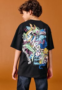 Black cotton t-shirt featuring colorful monster graphics on the back, including skulls and whimsical designs. Short sleeves and relaxed fit.