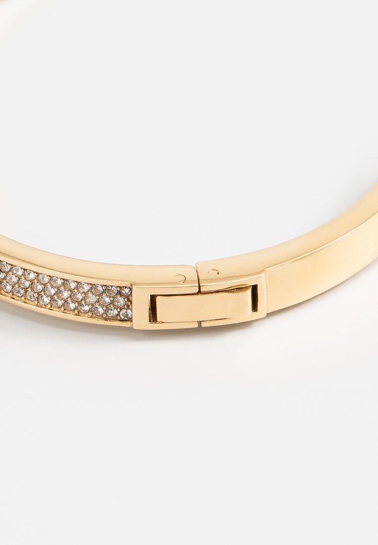 Bracelet doré avec une surface lisse et polie, ornée d'une rangée de cristaux clairs et scintillants au centre. Équipé d'un fermoir robuste.
