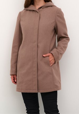 Kvinde iført en brun hættefrakke i mellemlængde med forlommer og sorte bukser foran en ensfarvet lys baggrund.