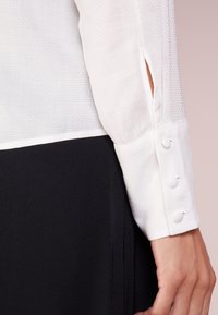 Chemise blanche en tissu texturé, à manches longues et avec des poignets boutonnés. Tissu noir visible en dessous, créant une apparence superposée.