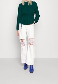 Blusa peplum verde scuro con maniche lunghe e vita con lacci, abbinata a jeans bianchi a gamba larga con ginocchia strappate e stivaletti blu.