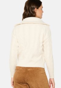 Kreminės spalvos megztinis su dideliu apykakle, su raštuotomis rankogaliais ir apačia. Dėvimi su rudais aksominiais kelnėmis.