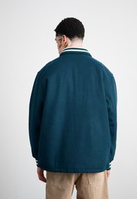 Teal fleecejakke med avslappet passform, ribbet krage og stripet detaljer ved mansjettene. Glatt tekstur med ensfarget stoff og minimale sømmer.