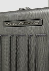 Harde koffer van gestructureerd donkergrijs materiaal met verticale groeven. Voorzien van een metalen logoplaatje, ritssluiting en strak design.