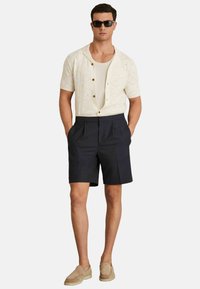 Cămașă tricotată cu mânecă scurtă, de culoare crem, cu un model hexagonal, asociată cu pantaloni scurți bleumarin. Pantofi slip-on bej și ochelari de soare închiși la culoare completează ținuta.