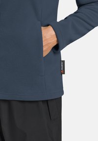 Blaue Fleecejacke mit glatter Textur und flachen Nähten, ausgestattet mit einer Seitentasche und einem schwarzen Polartec-Label am Saum.