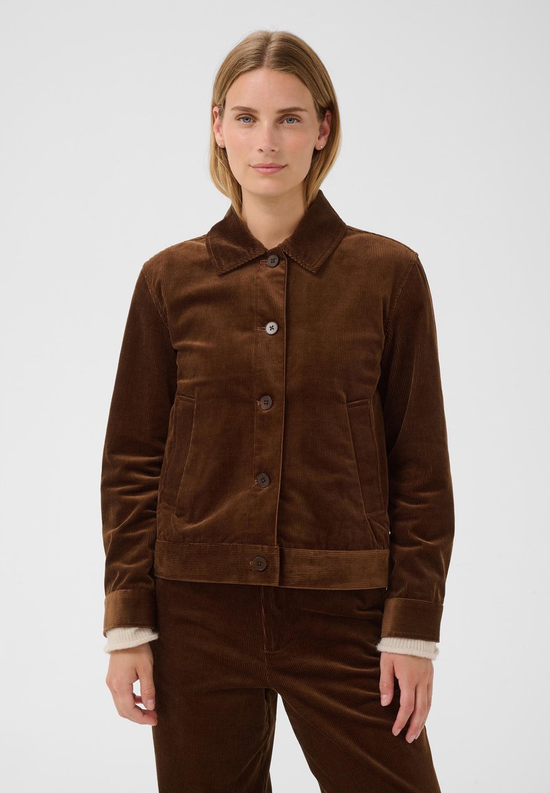 Femme portant une veste en velours côtelé marron avec des boutons et un pantalon assorti, debout devant un fond gris clair uni.