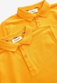 Dos camisetas polo amarillas de mezcla de algodón, con cuello, tres botones en la parte frontal y detalles cosidos. Etiquetas visibles en el interior de los cuellos.