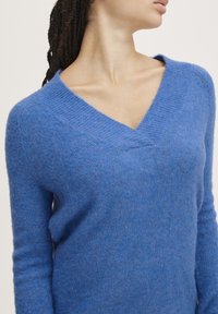 Zila adīta džempera ar V veida kaklu, izgatavota no mīksta, strukturēta materiāla. Rūpīgi izstrādātas garas piedurknes un brīvs piegriezums. Nav redzamu rakstu vai metāla detaļu.