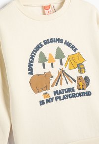 Sudadera de color crema con un gráfico de temática de campamento: oso, árboles, tienda de campaña y leña. El texto dice "La aventura comienza aquí, la naturaleza es mi patio de juegos."