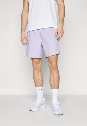 Homme portant un short de sport lilas, des chaussettes blanches, des baskets blanches et un débardeur blanc, debout devant un fond clair uni.