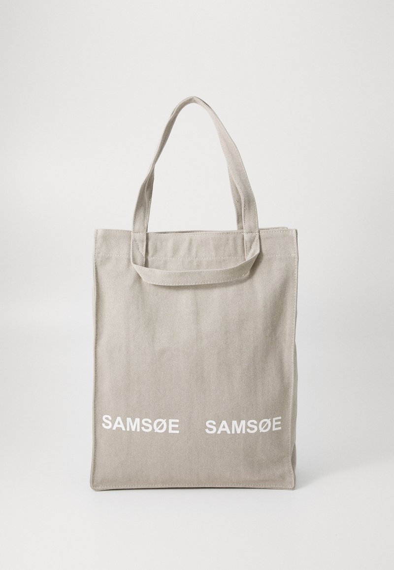 Pilkos kanvos tote maišelis su dviem rankenomis; priekyje yra balta spausdinta užrašas "SAM SøE"; minimalistinis dizainas, stačiakampio forma, minkšta tekstūra.