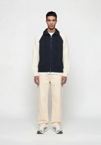 Άνδρας που φοράει μπλε σκούρο και άσπρο πλεκτό πουλόβερ με φερμουάρ, μπεζ παντελόνι και λευκά αθλητικά παπούτσια, στέκεται μπροστά σε ένα απλό λευκό φόντο.