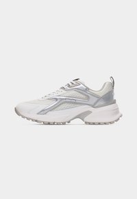 Baskets de sport blanches et argentées avec une semelle épaisse, des panneaux en mesh, des lacets et une languette à l'arrière sur un fond clair uni.