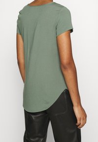 Zelený top s krátkým rukávem a zaobleným výstřihem, se zakřiveným lemem, vyrobený z jemné látky, v kombinaci s černými volnými kalhotami.