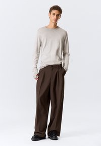 Chemise à manches longues gris clair associée à un pantalon brun foncé taille haute, plissé, complété par des chaussures noires. Design simple, coupe décontractée.
