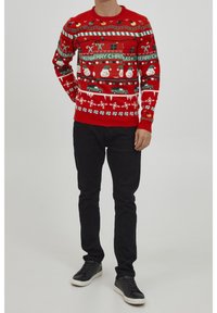 Roter gestrickter Weihnachtspullover mit festlichen Mustern, darunter Schneemänner, Autos und Schneeflocken. Getragen mit schwarzen Hosen und Sneakers.
