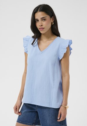 Kobieta w jasnoniebieskiej bluzce bez rękawów z dekoltem w serek i falbankami na ramionach oraz ciemnych szortach dżinsowych, uzupełniona o złote kolczyki koła i bransoletkę.