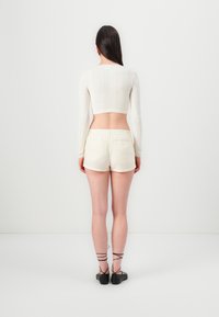Maglione bianco corto con motivo testurizzato, abbinato a pantaloni corti beige chiaro. La modella indossa scarpe nere con cinturini alla caviglia e ha lunghi capelli scuri.