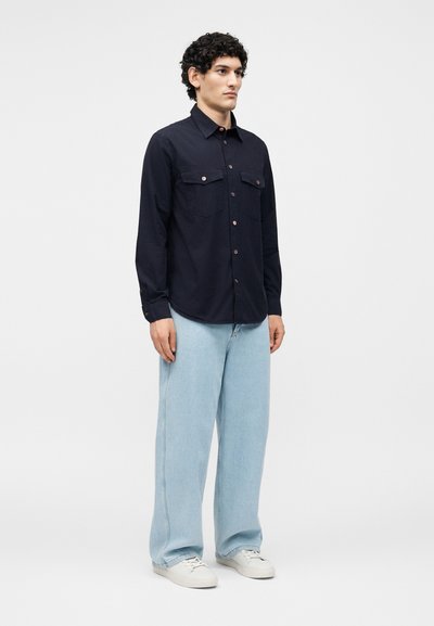 Άνδρας με σγουρά σκούρα μαλλιά, ντυμένος με μπλε πουκάμισο με κουμπιά, ανοιχτό μπλε φαρδιά τζιν και λευκά αθλητικά παπούτσια, στέκεται μπροστά σε έναν απλό φόντο.