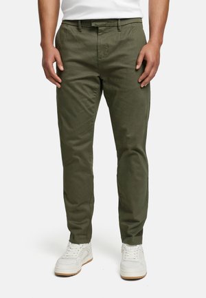 Mann iført olivengrønne slim fit-bukser og hvite joggesko, stående mot en ensfarget hvit bakgrunn.