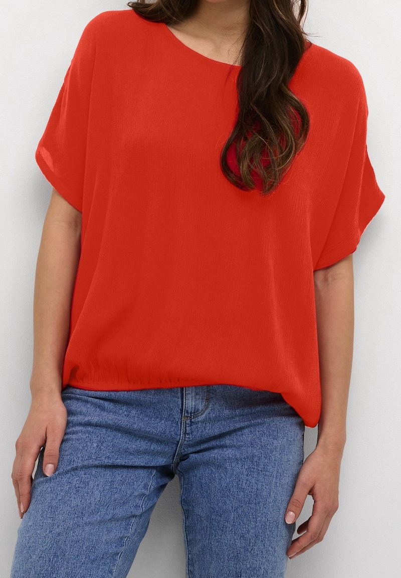 Blouse rouge à manches courtes avec un col rond et une coupe ample, associée à un jean en denim bleu clair. Texture lisse, design décontracté.